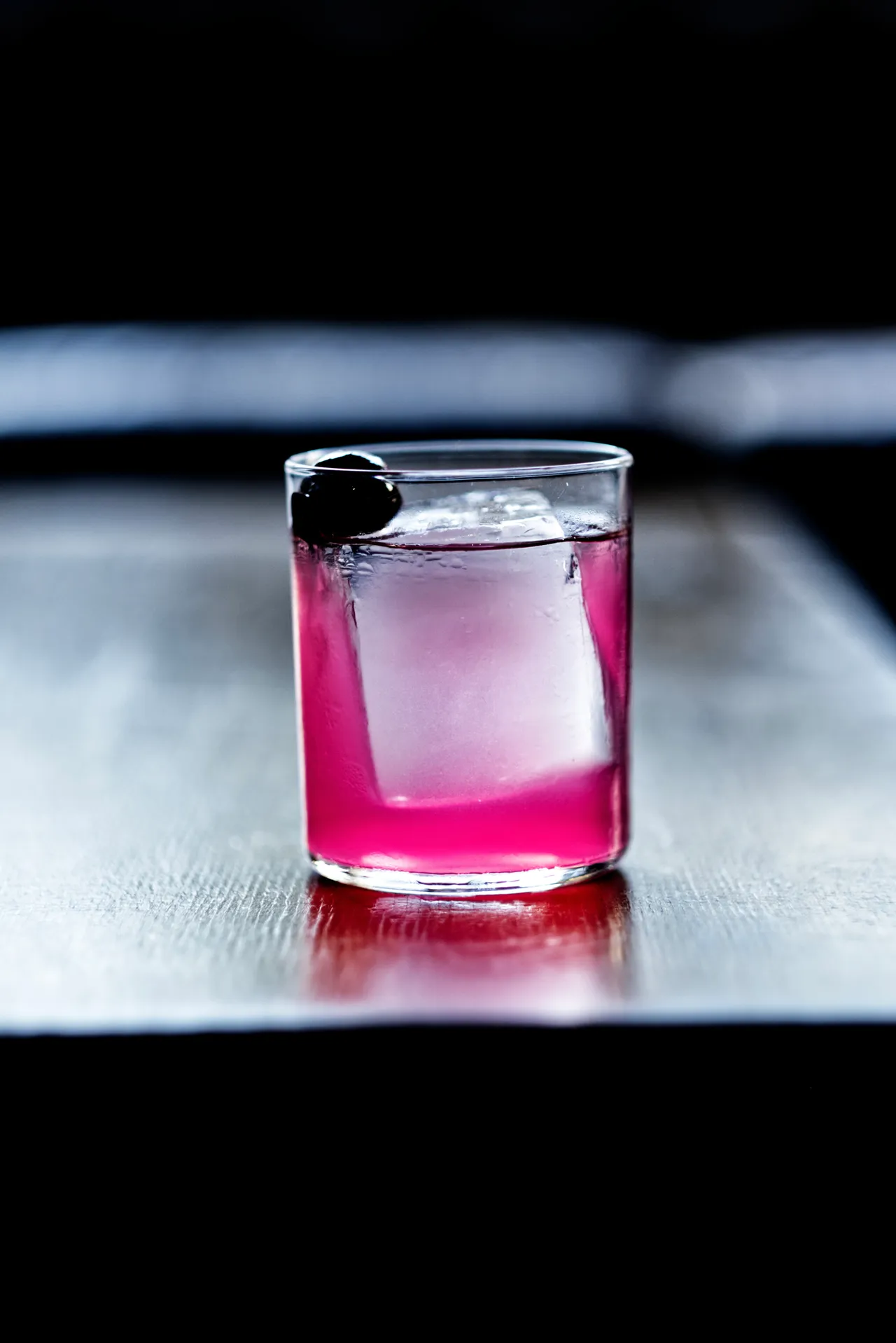 Cocktail magenta con ciliegia scura su un bancone lucido, riflesso rosa