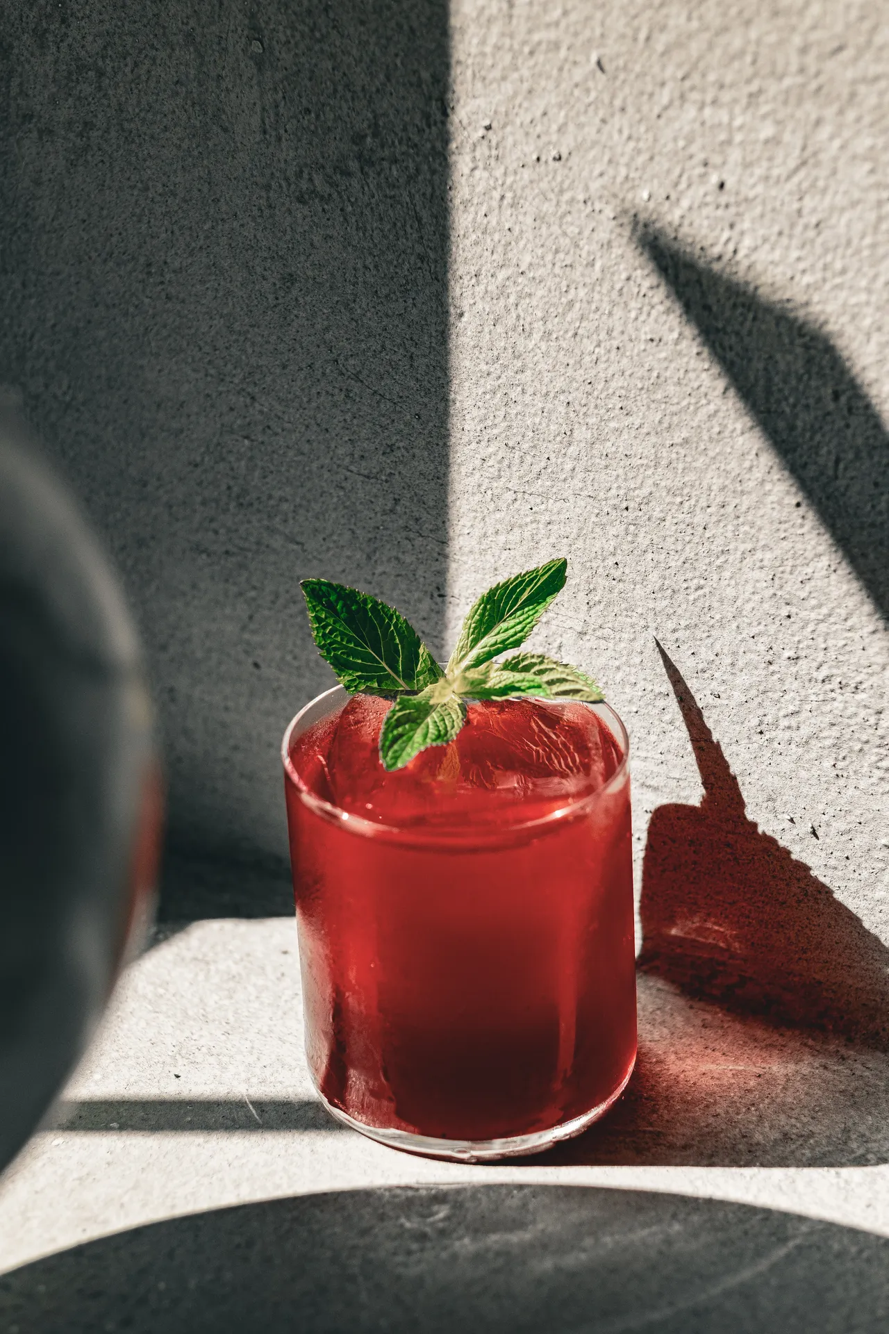 Cocktail rosso con foglie di menta in un tumbler, ombre nette su una parete di cemento