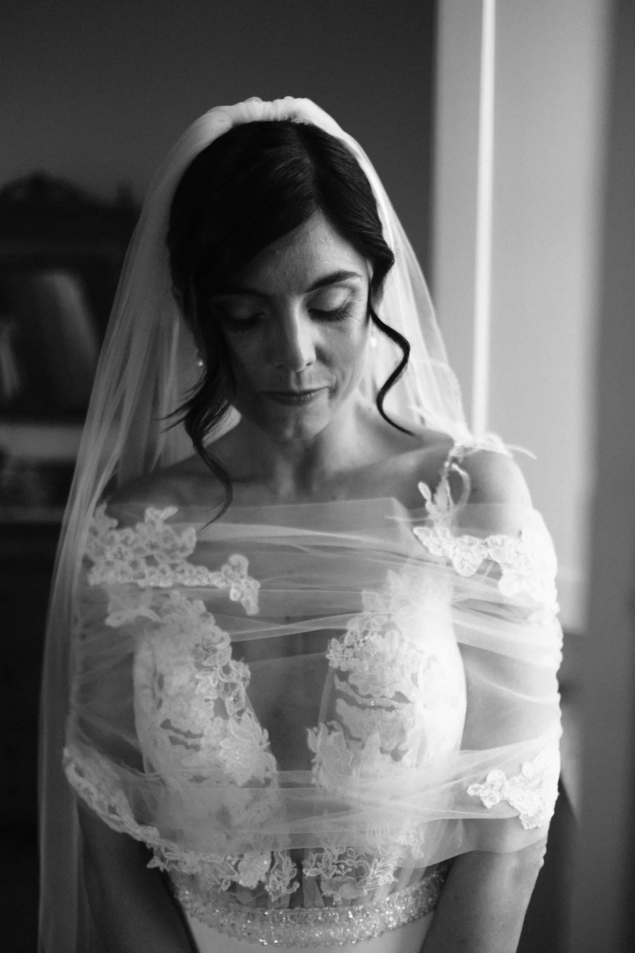 Sposa con velo e abito in pizzo accanto a una finestra, lo sguardo abbassato, in bianco e nero