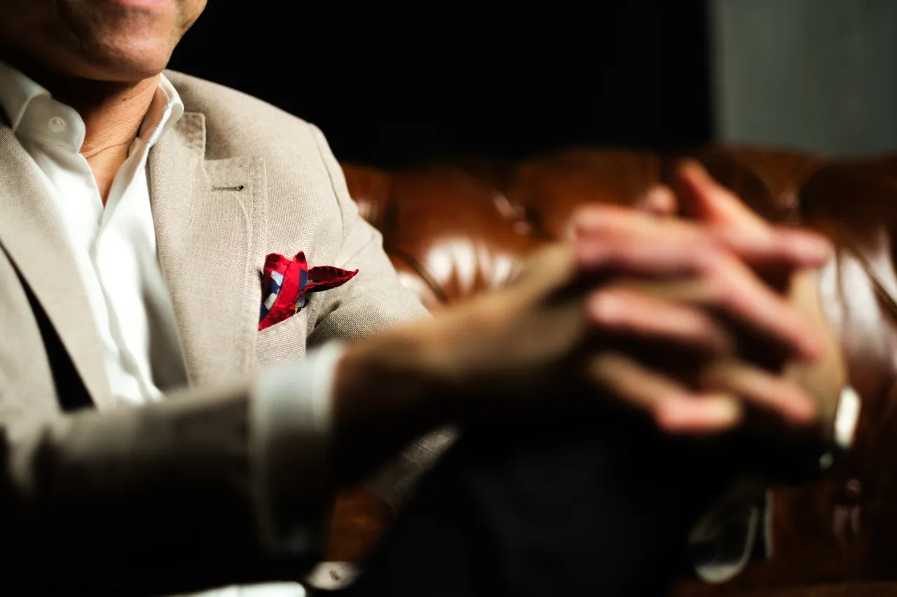 Dettaglio di mani intrecciate, giacca chiara con pochette rossa davanti a un divano chesterfield in pelle