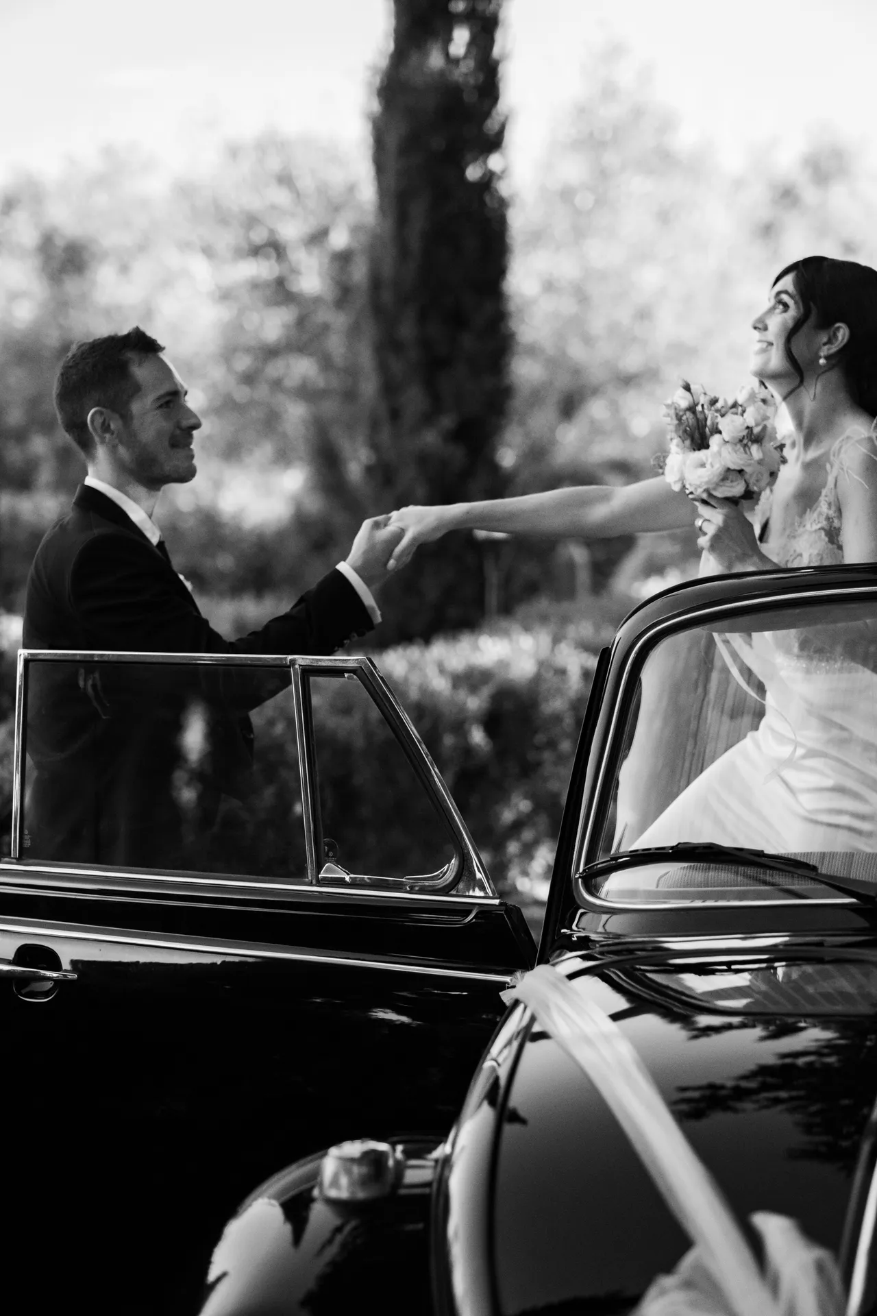 Lo sposo prende la mano della sposa che scende da una cabriolet d'epoca, tra i cipressi